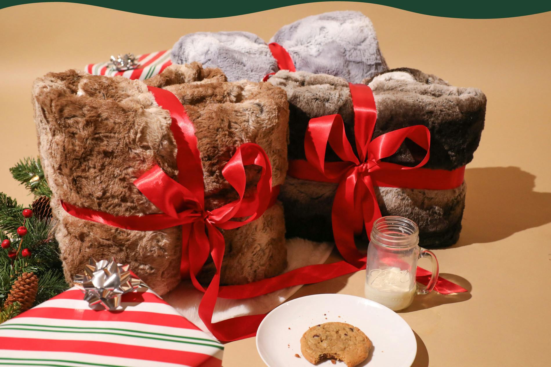 Photo of 3 different faux fur sherpa blankets (one brown, dark brown, and grey) wrapped in red ribbon like presents & sitting next to wrapped gift boxes, a plate of cookies & a glass of milk.