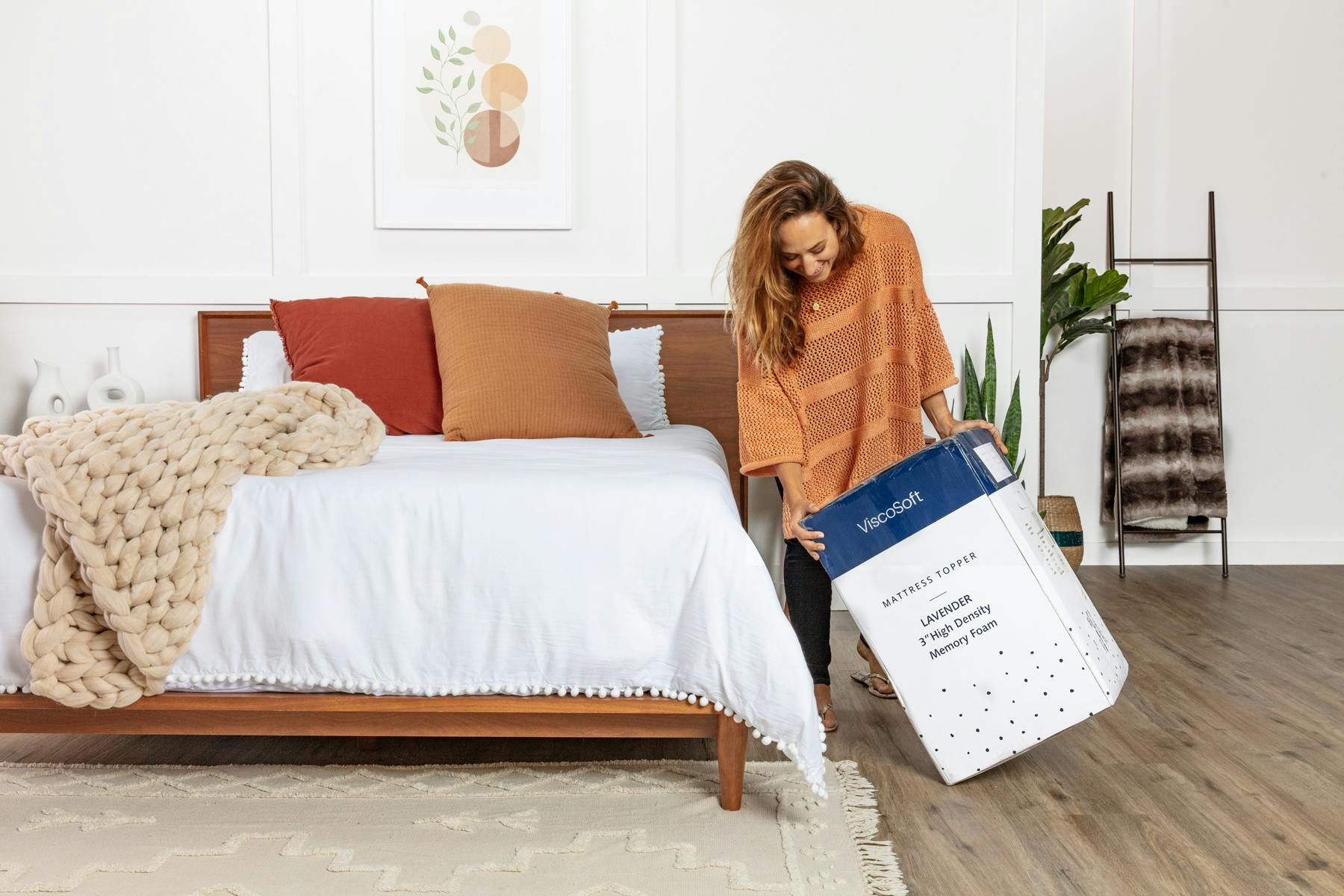 a woman standing beside a bed tilting a mattress topper box on it's side like she's about to open it