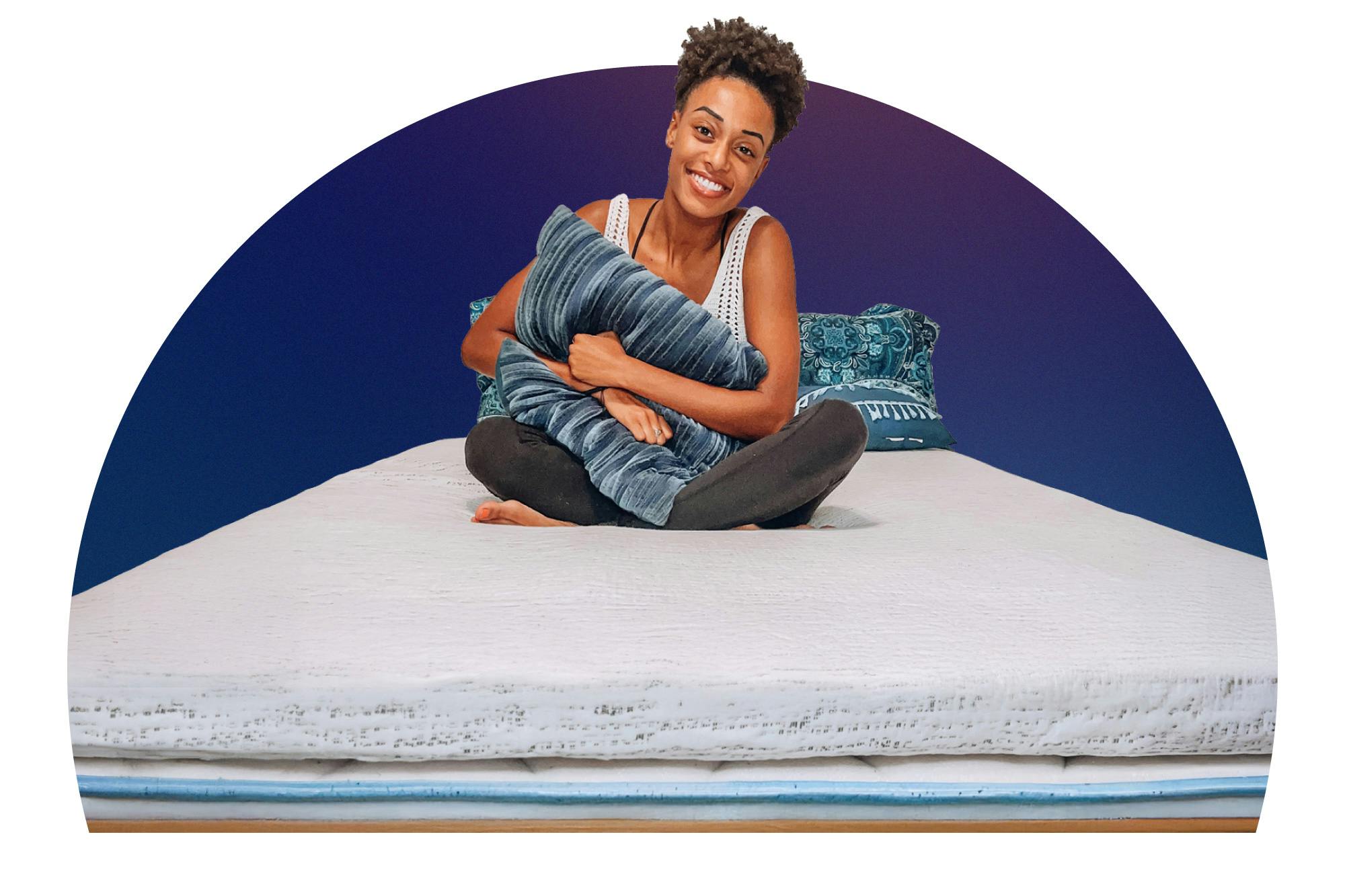 Woman sitting cross-legged on dorm bed, hugging a pillow, with a thick white mattress topper on her bed.