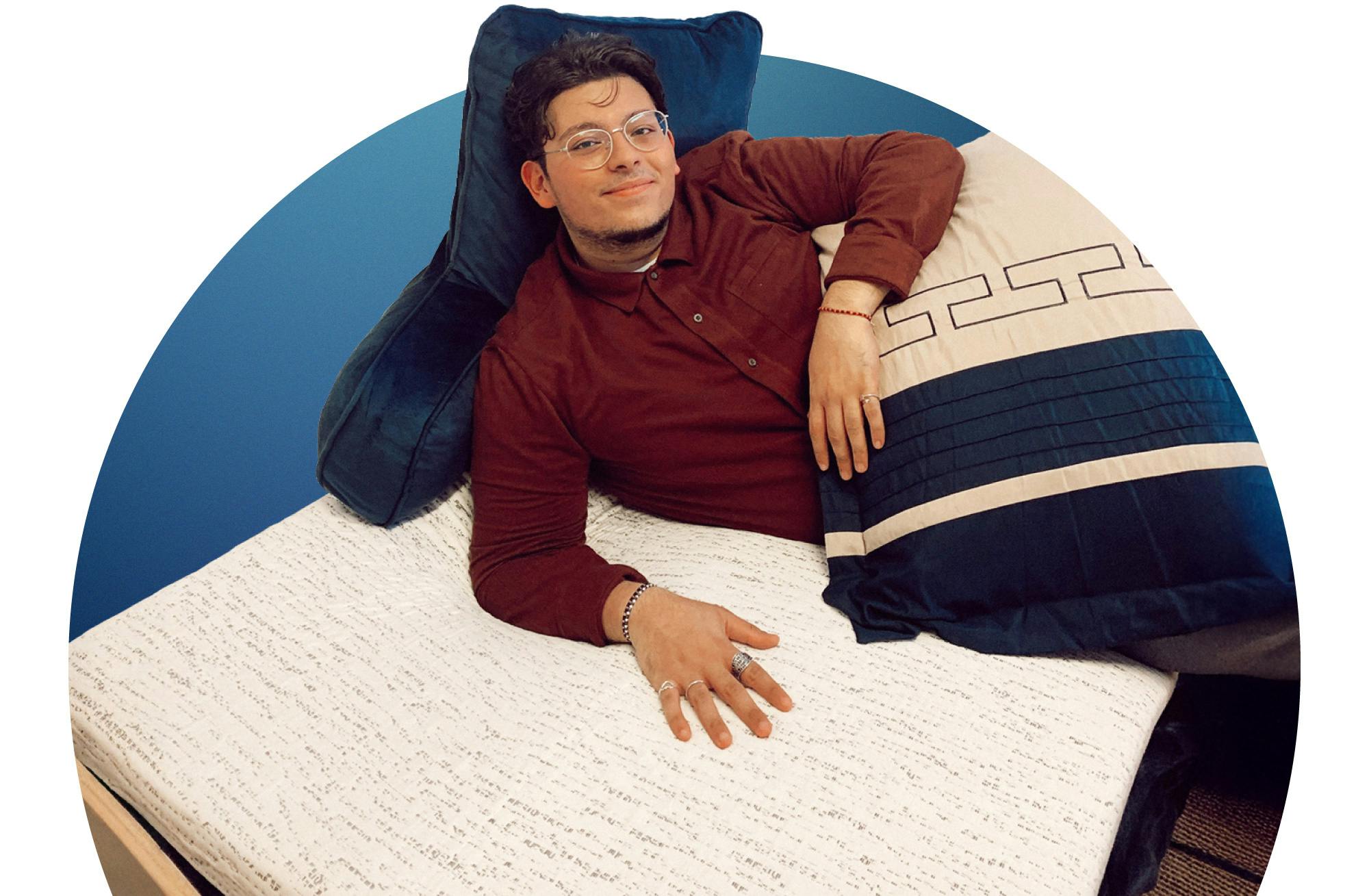Man lounging on college dorm bed with textured mattress topper and large pillows.