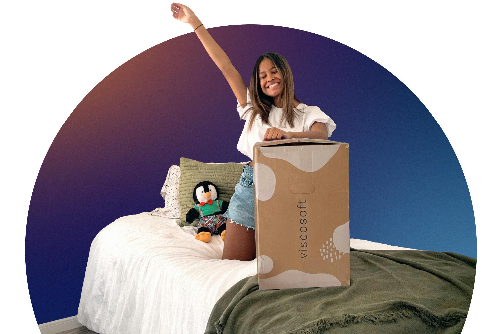 Smiling woman opening Viscosoft box on bed with stuffed penguin and pillows.