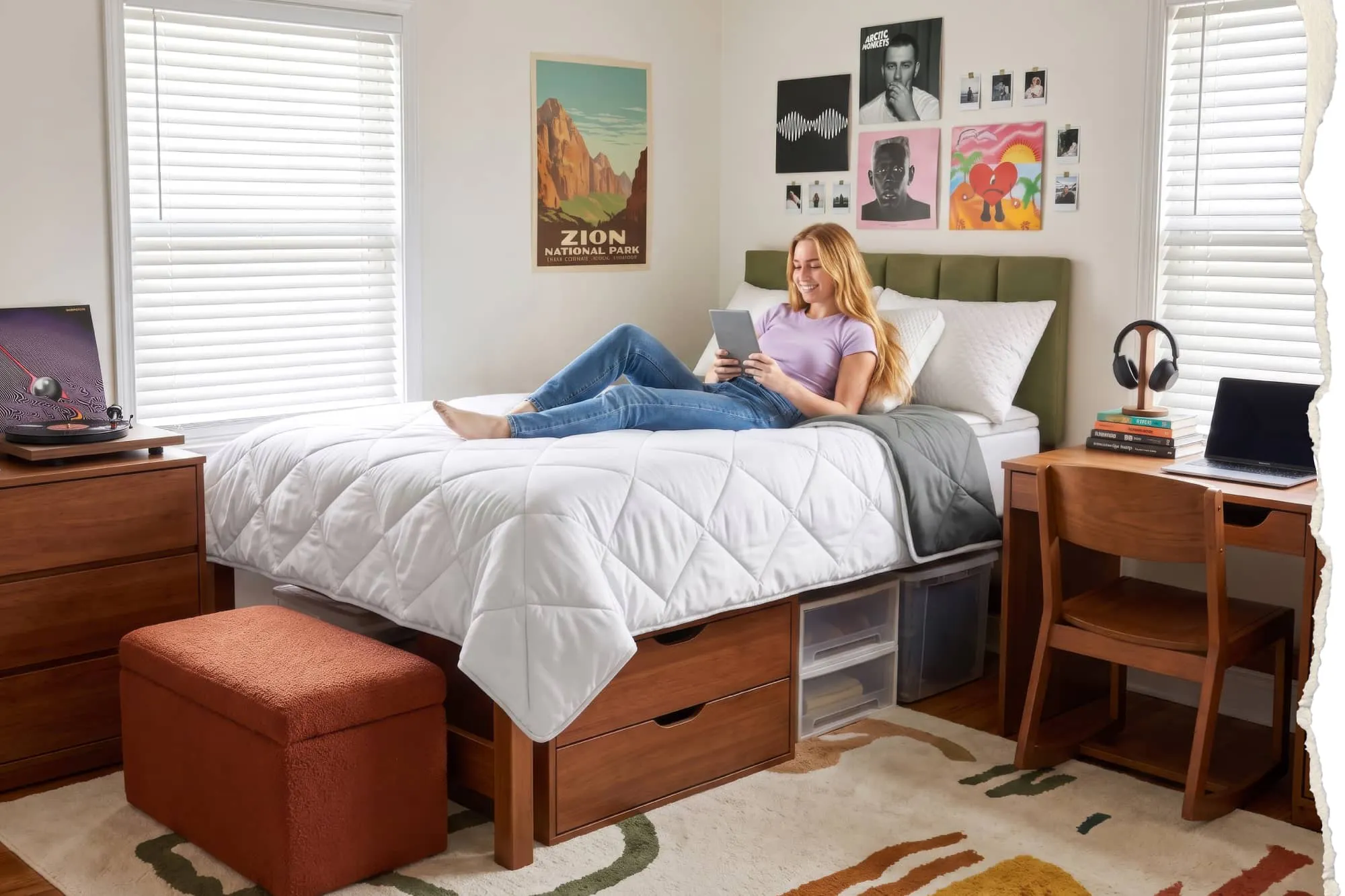 A college student lounging on their dorm bed fitted with a white pillow top mattress topper, a gray reversible comforter, and 3 plush white pillows.