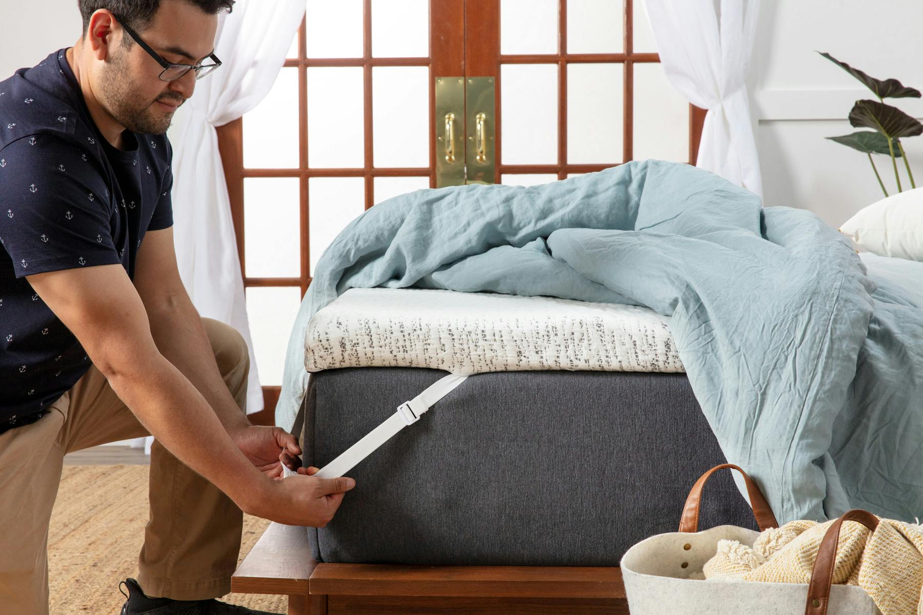 Man pulling white adjustable elastic strap of a thick white and blue mattress topper over the corner of a mattress. There is a blue comforter draped across the bed.
