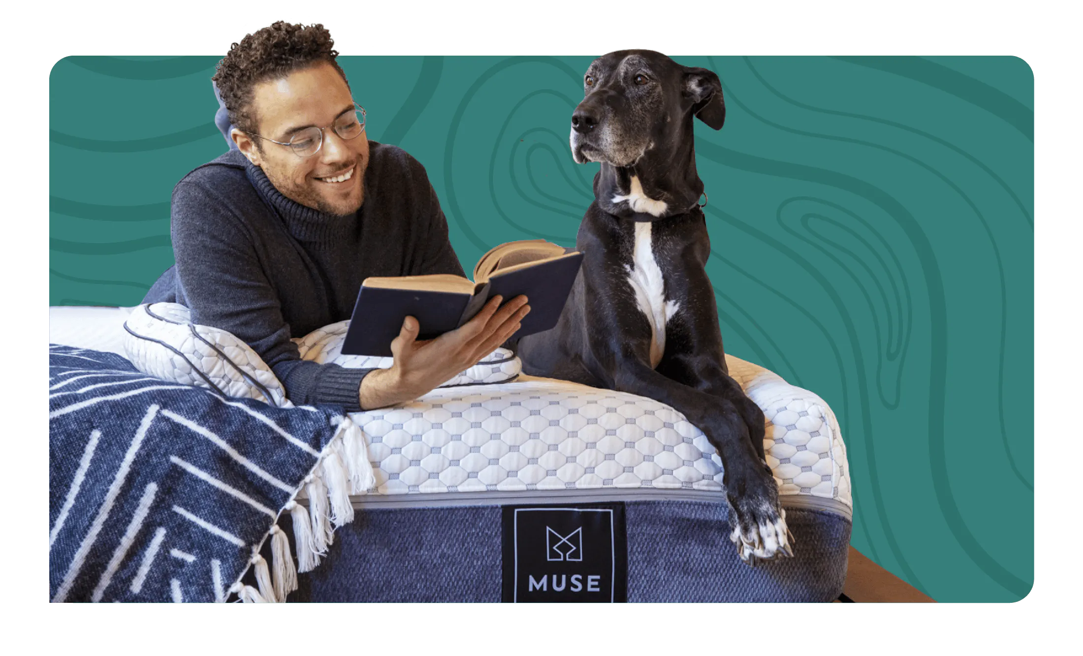 A man lounging while reading a book on a white and gray mattress with his gray Great Dane lays next to him