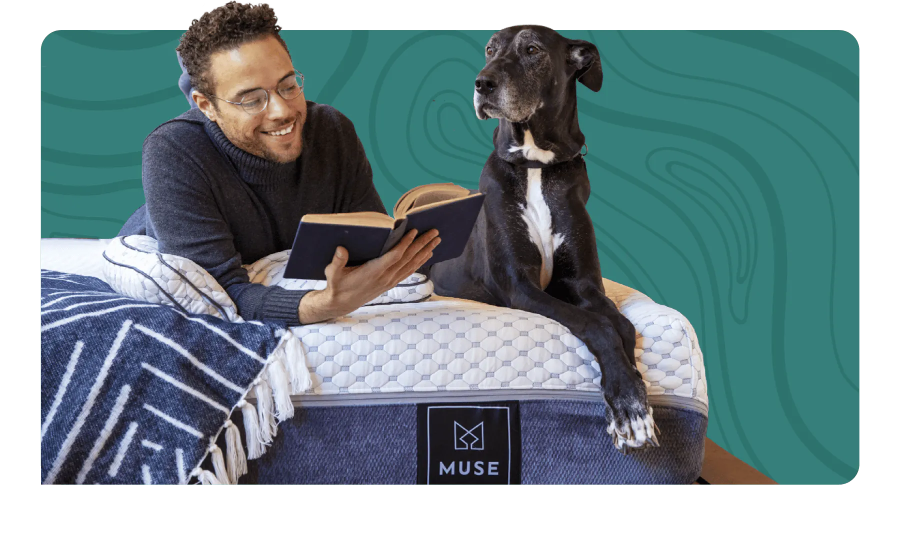A man lounging while reading a book on a white and gray mattress with his gray Great Dane lays next to him