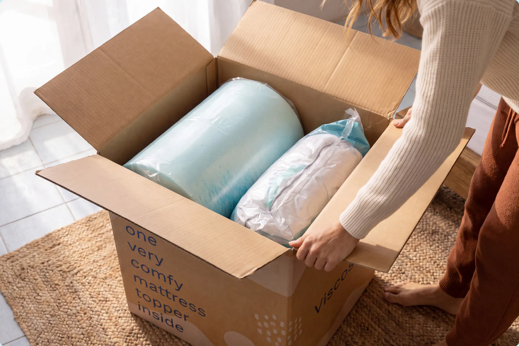 Woman opening a Viscosoft box holding a rolled blue gel memory foam topper and a vacuum-packed white quilted topper.